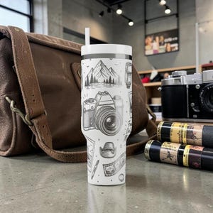 K&ouml;nnte beinhalten: Wei&szlig;er Reisebecher mit einer Schwarz-Wei&szlig;-Illustration einer Kamera, Bergen und anderen Elementen der Fotografie. Der Becher hat einen wei&szlig;en Deckel und einen Strohhalm. Eine braune Ledertasche und Vintage-Kameras befinden sich im Hintergrund.