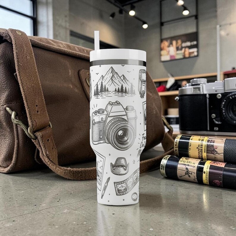 K&ouml;nnte beinhalten: Wei&szlig;er Reisebecher mit einer Schwarz-Wei&szlig;-Illustration einer Kamera, Bergen und anderen Elementen der Fotografie. Der Becher hat einen wei&szlig;en Deckel und einen Strohhalm. Eine braune Ledertasche und Vintage-Kameras befinden sich im Hintergrund.