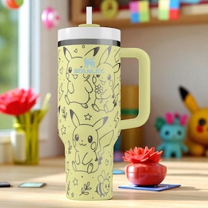 Puede incluir: Un vaso Stanley amarillo con tapa y asa blancas, con un diseño de Pikachu. El vaso tiene una pajita y está decorado con dibujos lineales negros de Pikachu, estrellas, corazones y rayos. El logotipo de Stanley es visible en la parte delantera.
