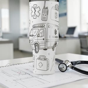 May include: A white tumbler featuring black line drawings of medical symbols, an ambulance, and a first aid kit. The tumbler is on a table with a stethoscope and medical paperwork.