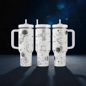 May include: Three white Stanley tumblers with handles and straws, featuring a black line art space theme. The design includes astronauts, planets, satellites, and stars. The Stanley logo is printed on the right tumbler. The tumblers are cylindrical.