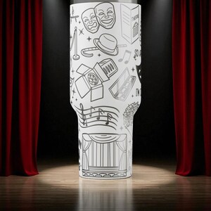 May include: A white tumbler with a black line drawing of theater-related elements, including masks, a stage, musical notes, and a spotlight. The tumbler is cylindrical with a slightly tapered base. The background features red curtains and a wooden stage.