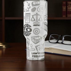 May include: A white tumbler with a black and white illustration of law-related icons and text. The tumbler features images of scales, money, and the text "BORN TO ARGUE" and "JUSTICE". The tumbler is on a dark wooden surface.