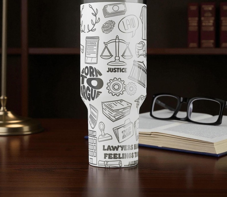 May include: A white tumbler with a black and white illustration of law-related icons and text. The tumbler features images of scales, money, and the text "BORN TO ARGUE" and "JUSTICE". The tumbler is on a dark wooden surface.