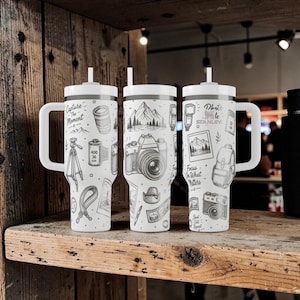 K&ouml;nnte beinhalten: Drei wei&szlig;e isolierte Becher mit Griffen und Strohhalmen, jeweils mit schwarz-wei&szlig;en Illustrationen zum Thema Fotografie. Die Designs umfassen Kameras, Berge, Filmrollen und Texte wie "Capture the Moment" und "Photo Life".
