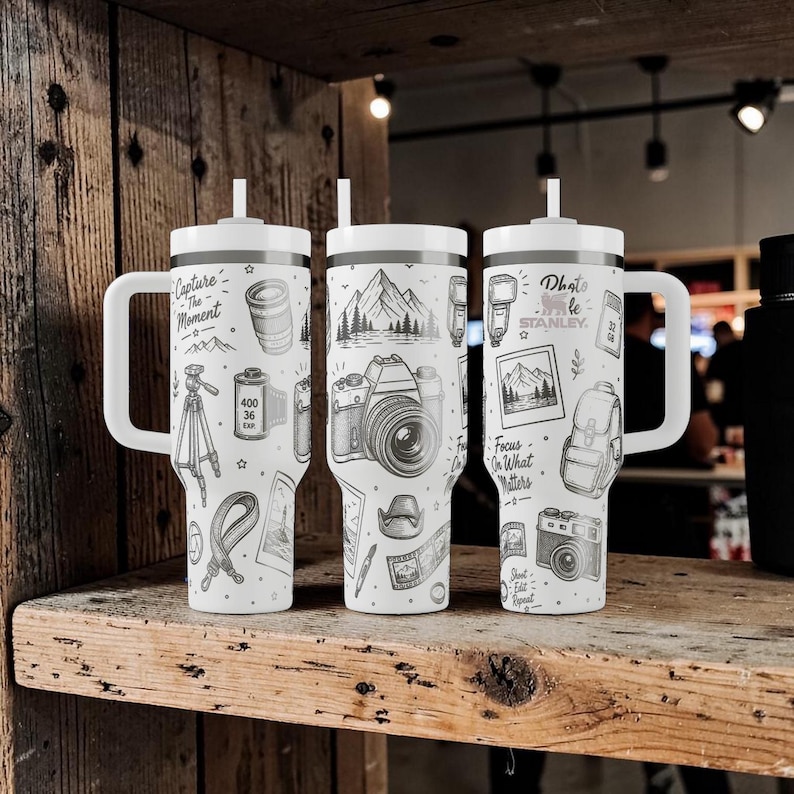 K&ouml;nnte beinhalten: Drei wei&szlig;e isolierte Becher mit Griffen und Strohhalmen, jeweils mit schwarz-wei&szlig;en Illustrationen zum Thema Fotografie. Die Designs umfassen Kameras, Berge, Filmrollen und Texte wie "Capture the Moment" und "Photo Life".