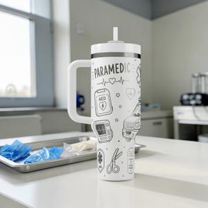 May include: White insulated tumbler with a handle and straw, featuring the word "PARAMEDIC" and medical-themed illustrations in black. Designs include a heartbeat, AED, ambulance, and scissors. The tumbler is on a white surface.