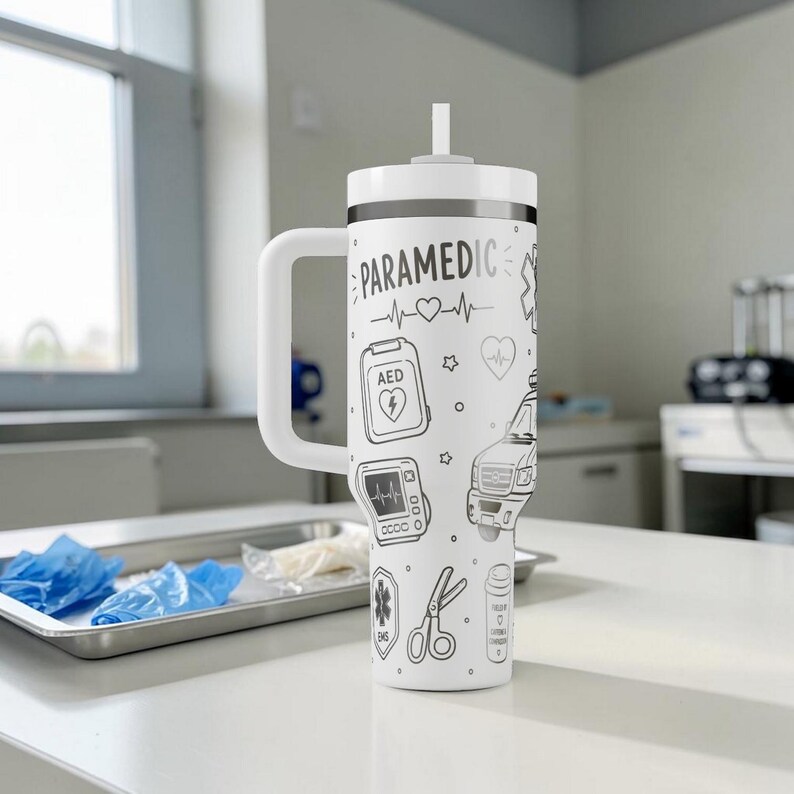May include: White insulated tumbler with a handle and straw, featuring the word "PARAMEDIC" and medical-themed illustrations in black. Designs include a heartbeat, AED, ambulance, and scissors. The tumbler is on a white surface.