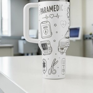 May include: White travel mug with a large handle, decorated with black line drawings of paramedic-related symbols, including an AED, ambulance, and heart rate monitor. The word "PARAMEDIC" is written at the top.