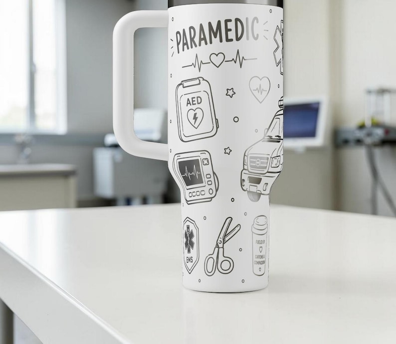 May include: White travel mug with a large handle, decorated with black line drawings of paramedic-related symbols, including an AED, ambulance, and heart rate monitor. The word "PARAMEDIC" is written at the top.