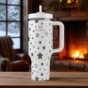 May include: A white Stanley tumbler with a handle and a straw, featuring a repeating pattern of grey stars of varying sizes across the exterior. The brand logo is printed in grey near the top rim of the cylindrical vessel, which has a tapered base.