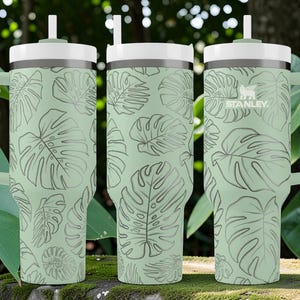 Puede incluir: Vaso Stanley verde claro con tapa y asa blancas, con un estampado repetido de hojas de monstera. El vaso tiene una pajita y el logotipo de Stanley. Adecuado para bebidas fr&iacute;as.