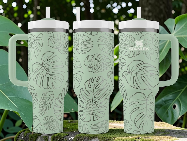 Puede incluir: Vaso Stanley verde claro con tapa y asa blancas, con un estampado repetido de hojas de monstera. El vaso tiene una pajita y el logotipo de Stanley. Adecuado para bebidas fr&iacute;as.
