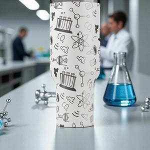 May include: A white tumbler with a science-themed pattern of atoms, beakers, and CO2 symbols. The tumbler is on a lab bench with scientific equipment and a flask of blue liquid. The background shows a laboratory setting.