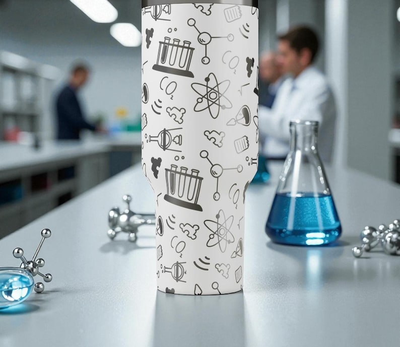 May include: A white tumbler with a science-themed pattern of atoms, beakers, and CO2 symbols. The tumbler is on a lab bench with scientific equipment and a flask of blue liquid. The background shows a laboratory setting.