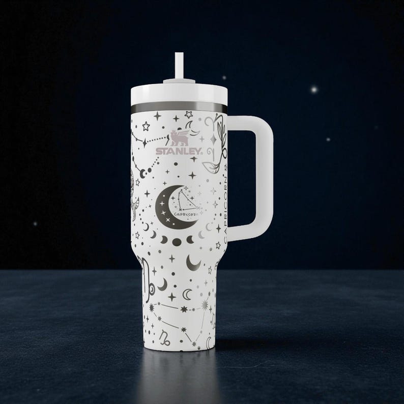 K&ouml;nnte beinhalten: Wei&szlig;er Stanley-Becher mit Griff und Strohhalm, mit einem Sternbild- und Monddesign. Der Becher tr&auml;gt das Wort "Capricorn" und das Stanley-Logo. Der Hintergrund ist dunkelblau mit Sternen.