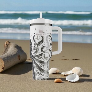 May include: White Stanley travel mug with a black octopus design. The mug has a white handle and lid with a straw. The design features an octopus with its tentacles wrapping around the mug. The mug is on a sandy beach with seashells.