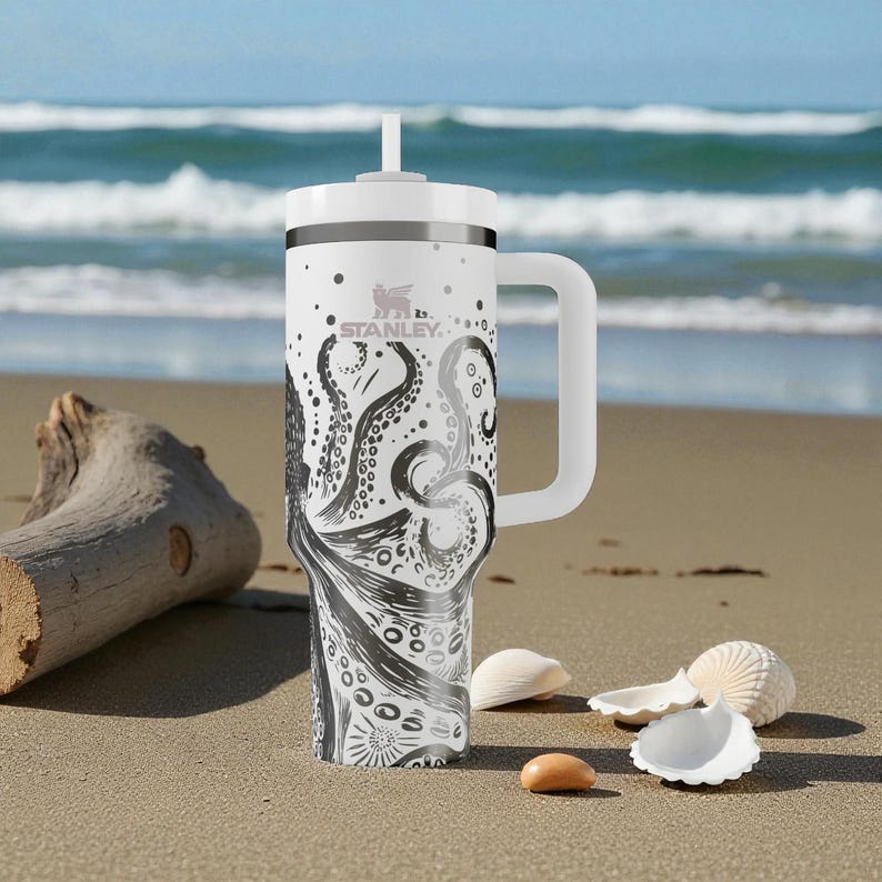 May include: White Stanley travel mug with a black octopus design. The mug has a white handle and lid with a straw. The design features an octopus with its tentacles wrapping around the mug. The mug is on a sandy beach with seashells.