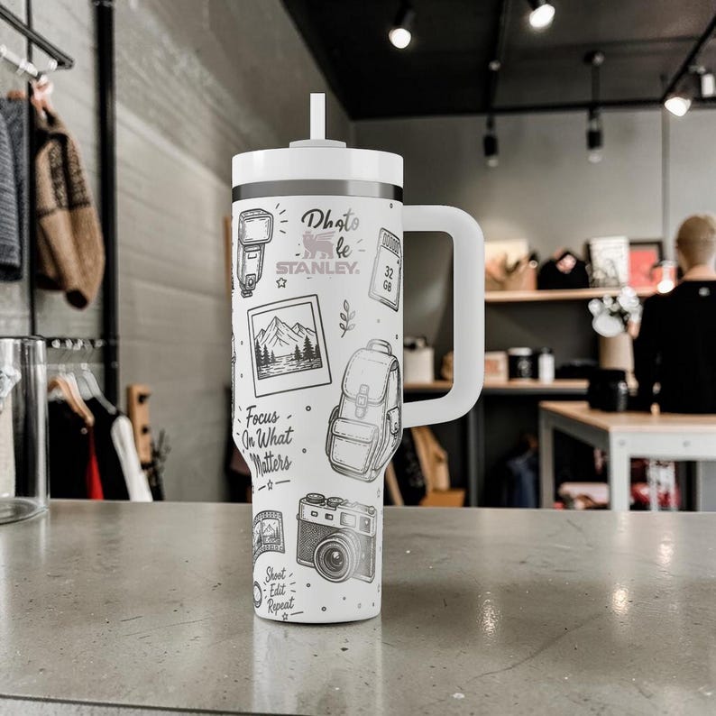K&ouml;nnte beinhalten: Wei&szlig;er isolierter Becher mit Henkel und Strohhalm, mit einem schwarz-wei&szlig;en Design zum Thema Fotografie. Das Design umfasst Illustrationen einer Kamera, eines Blitzes, eines Rucksacks und den Text "Photo by Stanley". Der Becher tr&auml;gt auch den Text "Focus on What Matters".