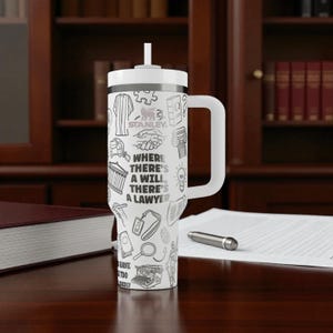 May include: White tumbler with a handle and a straw, featuring black line art of legal symbols and the text "WHERE THERE'S A WILL THERE'S A LAWYER." The tumbler is a Stanley brand and sits on a wooden desk.