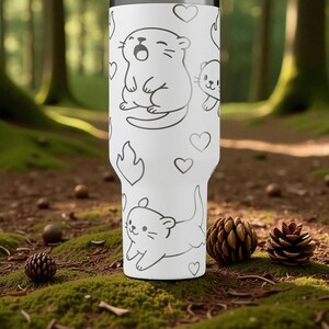 K&ouml;nnte beinhalten: Wei&szlig;er Becher mit einem sich wiederholenden Muster aus Cartoon-Ottern, Herzen und Flammen in Schwarz. Der Becher ist zylindrisch und scheint aus Edelstahl zu sein. Der Hintergrund ist ein Waldboden mit Moos, Tannenzapfen und Eicheln.