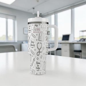 Puede incluir: Vaso Stanley blanco con asa y pajita, decorado con ilustraciones m&eacute;dicas en negro. El dise&ntilde;o incluye un estetoscopio, vendas y los textos "Stay Strong" y "Born to Help". El vaso est&aacute; sobre una superficie blanca.