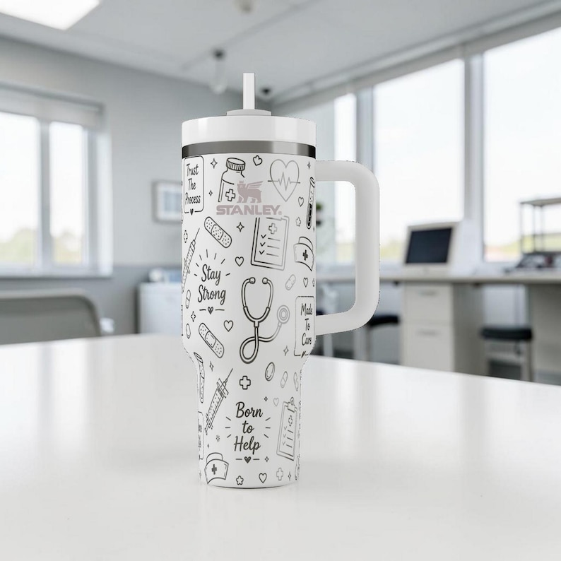 Puede incluir: Vaso Stanley blanco con asa y pajita, decorado con ilustraciones m&eacute;dicas en negro. El dise&ntilde;o incluye un estetoscopio, vendas y los textos "Stay Strong" y "Born to Help". El vaso est&aacute; sobre una superficie blanca.