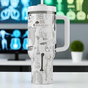 May include: A white Stanley tumbler with a handle and straw, featuring a black line art pattern of human skeletal anatomy, skulls, and radiation hazard symbols. The brand name Stanley is printed in black near the top of the cylindrical vessel.