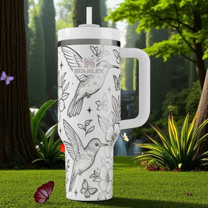 May include: White insulated tumbler with a handle, straw, and lid. The tumbler features a black and white hummingbird and floral design. The brand name "STANLEY" is printed on the side. The tumbler is designed for cold beverages.