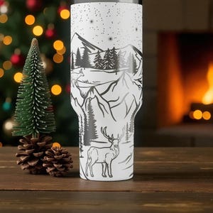 K&ouml;nnte beinhalten: Ein wei&szlig;er Becher mit einem schwarzen Berg- und Waldmotiv, das einen Hirsch und Sterne zeigt. Der Becher ist zylindrisch und scheint aus Edelstahl zu sein. Das Design umschlie&szlig;t den Becher und erzeugt eine malerische Landschaft.