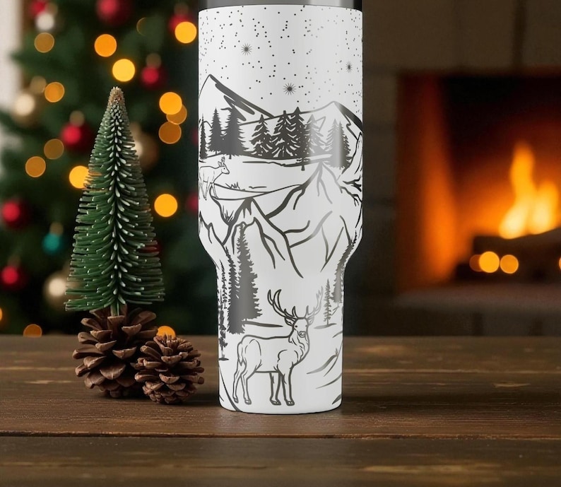 K&ouml;nnte beinhalten: Ein wei&szlig;er Becher mit einem schwarzen Berg- und Waldmotiv, das einen Hirsch und Sterne zeigt. Der Becher ist zylindrisch und scheint aus Edelstahl zu sein. Das Design umschlie&szlig;t den Becher und erzeugt eine malerische Landschaft.