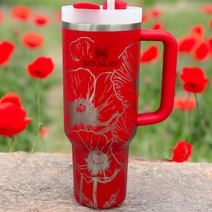 May include: A red Stanley tumbler with a handle, featuring a white lid and a straw. The exterior is decorated with an etched floral pattern of poppies. The Stanley logo is visible near the top center of the cup, positioned above the flower design.