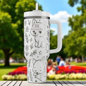 May include: White Stanley tumbler with a handle and straw. The tumbler features a black and white illustration of two foxes, flowers, and leaves. The Stanley logo is visible. The tumbler is sitting on a wooden surface.