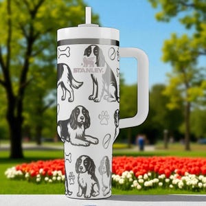 May include: A white Stanley tumbler with a handle and straw, featuring a repeating black and white graphic pattern of various spaniel dogs, paw prints, bones, and tennis balls. The Stanley logo is printed in grey on the upper front section.