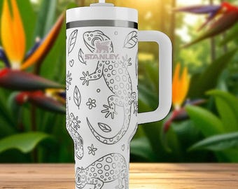 Puede incluir: Un vaso Stanley blanco con asa y pajita, que presenta un patrón repetitivo de líneas negras de geckos, hojas y pequeñas flores. El logotipo de Stanley está impreso en negro en la parte superior frontal del recipiente cilíndrico para bebidas.