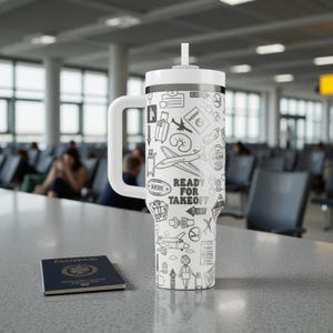 May include: A white travel mug with a handle and a straw, decorated with black travel-themed illustrations and the text "READY FOR TAKEOFF." A passport is on the table. The mug is on a table in an airport.