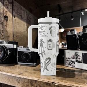 K&ouml;nnte beinhalten: Wei&szlig;er Reisebecher mit Henkel und Strohhalm, mit einem schwarz-wei&szlig;en Design zum Thema Fotografie. Der Becher ist mit Illustrationen eines Kameraobjektivs, einer Filmrolle, eines Stativs und dem Text "Capture The Moment" verziert.