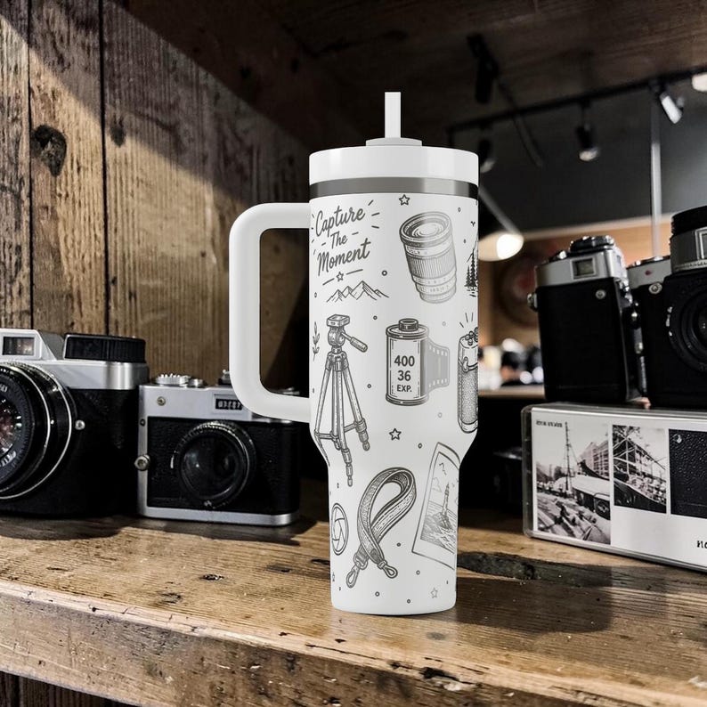 K&ouml;nnte beinhalten: Wei&szlig;er Reisebecher mit Henkel und Strohhalm, mit einem schwarz-wei&szlig;en Design zum Thema Fotografie. Der Becher ist mit Illustrationen eines Kameraobjektivs, einer Filmrolle, eines Stativs und dem Text "Capture The Moment" verziert.