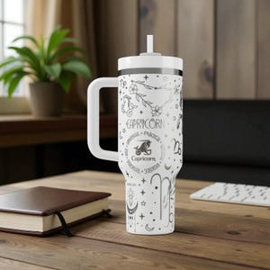 K&ouml;nnte beinhalten: Wei&szlig;er Becher mit Henkel, Strohhalm und einem schwarz-wei&szlig;en Steinbock-Tierkreiszeichen. Das Design beinhaltet das Wort "Steinbock", eine Ziegenillustration und andere Tierkreiszeichen. Der Becher steht auf einer Holzoberfl&auml;che.