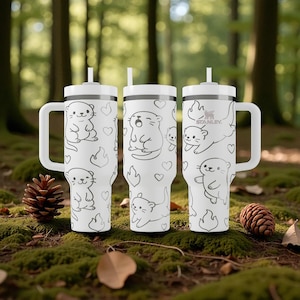 K&ouml;nnte beinhalten: Drei wei&szlig;e isolierte Becher mit Otter-Illustrationen, Herzen und Flammen. Jeder Becher hat einen wei&szlig;en Griff, Deckel und Strohhalm. Das Stanley-Logo ist auf einem Becher sichtbar. Auf einer moosigen Oberfl&auml;che.