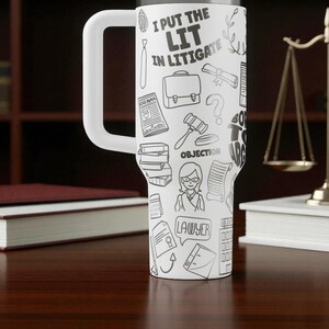 May include: White travel mug with a large handle, covered in black and white lawyer-themed doodles and the text "I PUT THE LIT IN LITIGATE." The mug is cylindrical and sits on a wooden surface.
