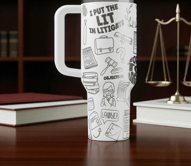 May include: White travel mug with a large handle, covered in black and white lawyer-themed doodles and the text "I PUT THE LIT IN LITIGATE." The mug is cylindrical and sits on a wooden surface.