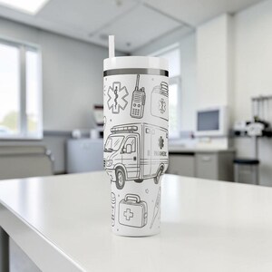 May include: White tumbler with a black and white paramedic-themed design. The design includes an ambulance, medical symbols, a radio, and a first aid kit. The tumbler has a white straw and a black band around the top.