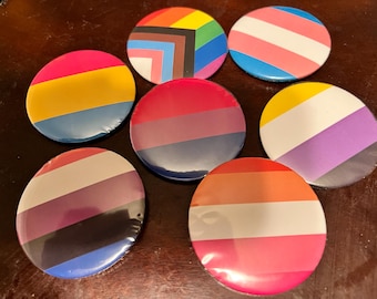 Puede incluir: Siete botones redondos con diferentes diseños de banderas del orgullo. Los botones son todos del mismo tamaño y están dispuestos en un círculo. Las banderas incluyen la bandera transgénero, la bandera bisexual, la bandera lesbiana, la bandera pansexual, la bandera genderfluid, la bandera no binaria y la bandera asexual.