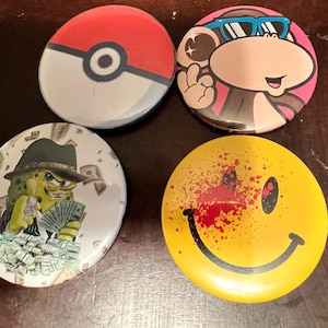 May include: Four round button pins with different designs. The first pin features a red and white Pokeball design. The second pin features a cartoon monkey wearing sunglasses. The third pin features a green cartoon character with money. The fourth pin features a yellow smiley face with red splatters.