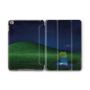 Puede incluir: Funda de tableta abierta con un paisaje sereno: una colina verde bajo un cielo estrellado. Una rana de dibujos animados con camisa azul y pantalón blanco meditando sobre una esterilla azul. La funda es blanca.