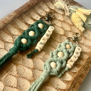 Peut inclure: Deux porte-clés en macramé verts, avec des perles en bois et les mots "PEACE" et "HOPE". Les porte-clés ont un design tressé et sont attachés à des fermoirs métalliques. Ils sont présentés sur un plateau en bois sculpté.