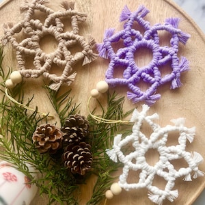 Peut inclure: Trois décorations de Noël en macramé en beige, violet et blanc, disposées sur un plateau en bois avec des pommes de pin et du feuillage. Chaque flocon de neige a un centre circulaire et des bords frangés, avec des perles en bois. Décoration de Noël artisanale.