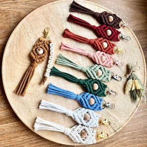 Peut inclure: Une collection de 12 porte-clés en macramé faits à la main avec des perles en bois et des glands de différentes couleurs, notamment le marron, le rouge, l'orange, le rose, le vert, le bleu et le blanc. Les porte-clés sont disposés sur un plateau en bois avec le mot "MARRAKECH" orthographié en lettres blanches.