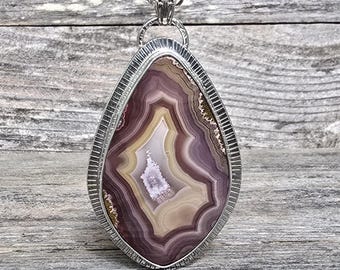 Puede incluir: Un collar con colgante en forma de lágrima con una piedra de ágata pulida en tonos morados, blancos y beige. La piedra está engastada en un marco de metal plateado texturizado y cuelga de una delicada cadena.